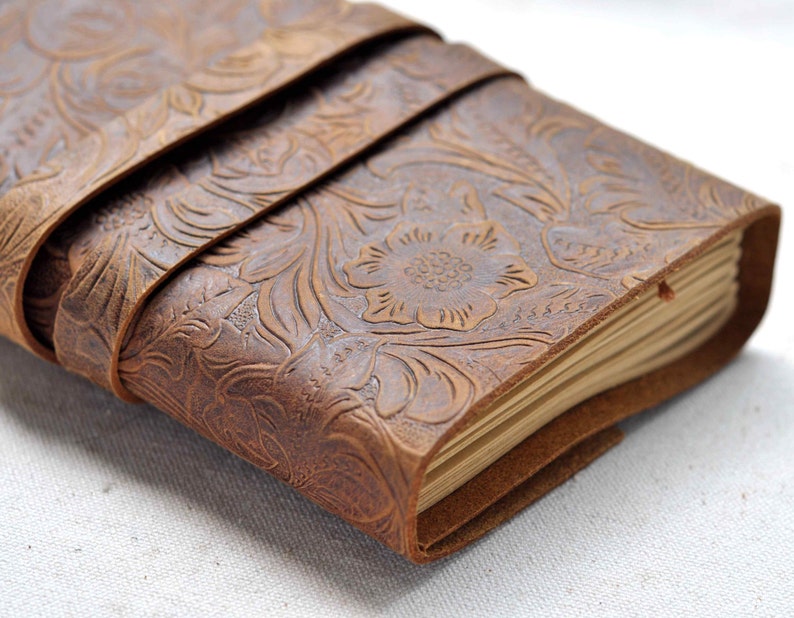 May include: A brown leather journal with a floral embossed design. The journal is closed and has a leather strap wrapped around it.
