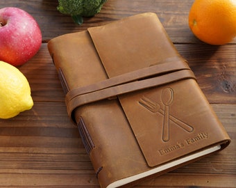 May include: A brown leather-bound journal with a strap closure. The cover has an embossed design of a fork and spoon crossed, and the text "Emma's Family".