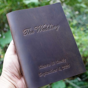 Può includere: Un diario in pelle marrone scuro con il testo in rilievo "The Wedding". Sotto, il diario è personalizzato con i nomi "Shawn & Sandy" e la data "6 settembre 2025."