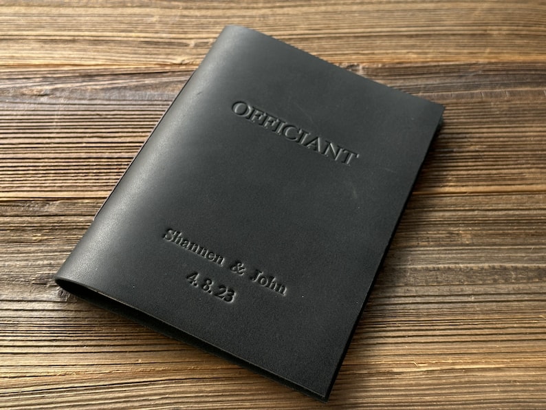 May include: Black leather officiant book with the text "Officiant" embossed on the front cover. The names "Shannon & John" and the date "4.8.23" are also embossed on the cover.