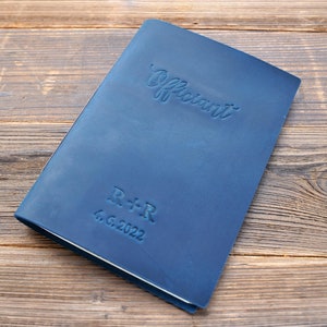 May include: A dark blue leather-bound journal with the word "Officiant" embossed on the front cover. The journal also has the initials "R+R" and the date "4.6.2022" embossed on the front cover.