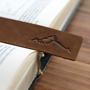 May include: A brown leather bookmark with a mountain range design embossed on it. The bookmark is placed in a book.
