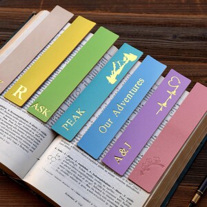 Può includere: Sette segnalibri in pelle colorati con lettere e disegni dorati. I segnalibri sono disposti in fila su un libro aperto. I disegni includono le lettere "AK", "R", "ASK", "PEAK", "Our Adventures", "A&J" e un cuore con una linea di battito cardiaco.