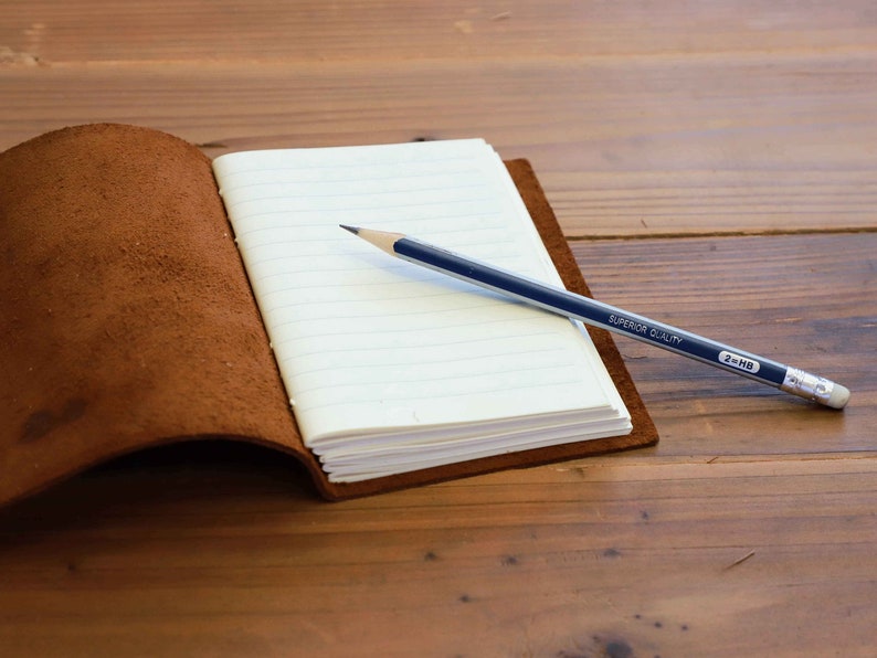 Pu&ograve; includere: Un diario rilegato in pelle marrone con una pagina bianca aperta e una matita appoggiata sulla pagina. La matita &egrave; blu e ha la scritta "SUPERIOR QUALITY 2HB" stampata su di essa.