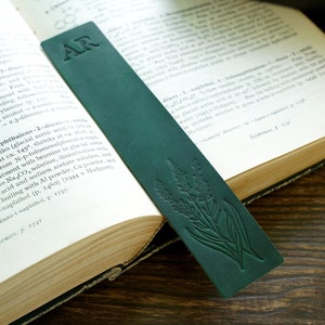 Może przedstawiać: Zakładka skórzana w ciemnozielonym kolorze z wytłoczonymi inicjałami "AR" z przodu. Zakładka ma kwiatowy wzór z kępą kwiatów lawendy.