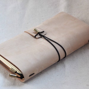 May include: A light brown leather journal with a black elastic closure and brass accents. The journal is closed and lying on a white fabric surface.
