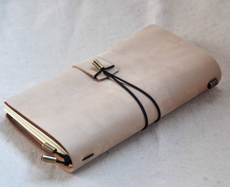 May include: A light brown leather journal with a black elastic closure and brass accents. The journal is closed and lying on a white fabric surface.