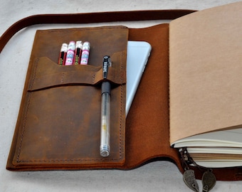 May include: A brown leather notebook with a pen holder, a pocket for pencils, and a strap. The notebook is open to reveal blank pages and a phone inside the cover. The notebook has a charm with the text "Don't forget me" on it.