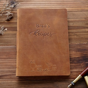 May include: A brown leather recipe book with the embossed text "Brad's Recipes." The cover features illustrations of cocktails. A pen rests on a wooden surface next to the book.