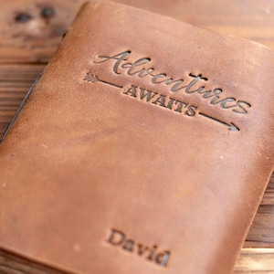 Pu&ograve; includere: Un diario in pelle marrone con il testo "Adventures Awaits" e una freccia sul davanti. Il nome "David" &egrave; inciso sul fondo della copertina.