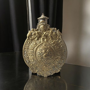 May include: A gold-coloured metal bottle with intricate carvings and a decorative lid. The bottle is sitting on a black surface.