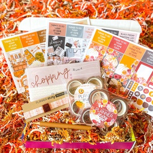 Puede incluir: Una caja rosa y marrón llena de suministros de planificación de otoño. La caja contiene pegatinas, cinta washi, un bolígrafo y un inserto de planificador. La caja está rodeada de papel de seda naranja y marrón.