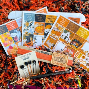 May include: A Halloween-themed planner box subscription with orange and black paper shred. The box includes stickers, a pen, a hair tie, and planner clips. The stickers feature pumpkins, cats, houses, and fall leaves. The hair tie says "#pumpkin everything".