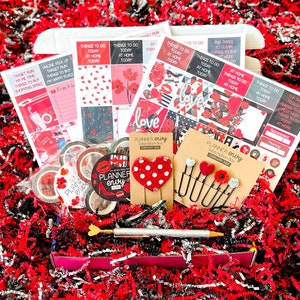 May include: A red and black planner box subscription box with the text "Planner Envy" and "February 2022". The box contains a variety of planner accessories, including stickers, washi tape, and a heart-shaped paper clip.