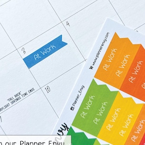 May include: A close-up of a planner page with the month of December 2023 visible. A blue banner sticker with the text "At Work" is on the 3rd. Colorful "At Work" stickers are shown. Text on the image says "Planner Envy".