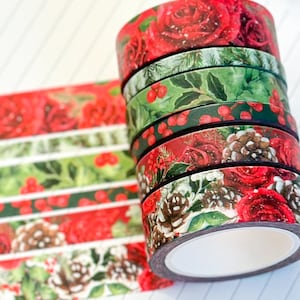 Puede incluir: Varios rollos de cinta adhesiva decorativa con diseños navideños. Los rollos muestran patrones de rosas rojas, acebo verde con bayas rojas y piñas. La cinta está dispuesta sobre una superficie blanca con un sutil patrón de rayas.