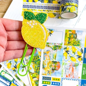 May include: A yellow lemon-shaped paper clip with green leaves is held in front of a planner with stickers and decorative tape. The stickers and tape feature lemon and blue patterns. The planner has the word "FRIDAY" printed on it.