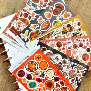 May include: A collection of planner stickers with a fall theme. The stickers feature images of pumpkins, turkeys, fall leaves, and other autumn-related graphics. The stickers are arranged on a wooden surface.