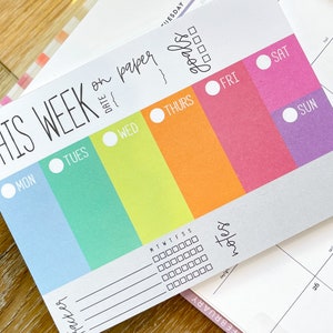 May include: A weekly planner with a colorful rainbow design. The planner has a section for each day of the week, with a space for notes and goals. The text "This Week on Paper" is at the top of the planner.