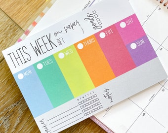 May include: A weekly planner with a colorful rainbow design. The planner has a section for each day of the week, with a space for notes and goals. The text "This Week on Paper" is at the top of the planner.