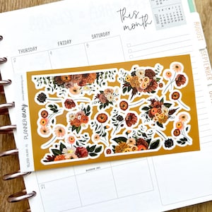 May include: A sheet of floral planner stickers in shades of orange, red, and cream, with green leaves, rests on an open planner. The planner has a white background with the text "This month" and a calendar.