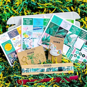 May include: A summer-themed planner subscription box with a unicorn bookmark, palm tree and monstera leaf paper clips, and stickers. The box includes the text "Planner Envy Subscription Box August 2021".