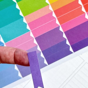 May include: A close-up of a colorful planner sticker sheet with a purple sticker being held by a finger. The sheet features a rainbow gradient of colors, including green, blue, pink, and red. The stickers are rectangular with a pointed bottom.
