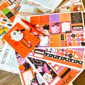 May include: A collection of Halloween-themed planner stickers, notebooks, and a pen. The stickers feature ghosts, pumpkins, and the word "BOO". The color palette includes orange, black, white, and purple. A notebook with the text "PLANNER envy" is also visible.