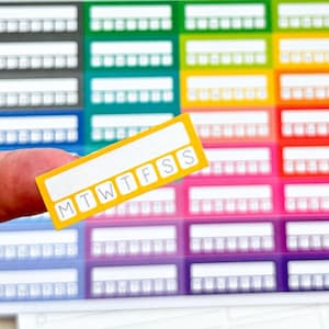 Puede incluir: Primer plano de una pegatina amarilla para planificador con los días de la semana impresos. La pegatina se sostiene delante de una hoja de pegatinas de planificador de colores. Los días de la semana están abreviados como MTWTFSS.