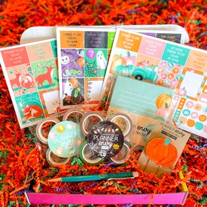 May include: A pink box filled with Halloween-themed planner supplies, including stickers, washi tape, a pen, and a pumpkin-shaped paper clip. The box is surrounded by orange and green shredded paper.