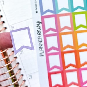 May include: A planner with a copper spiral binding and a sheet of colourful flag-shaped stickers. One sticker is held by a finger. The stickers are in shades of purple, pink, orange, green, and blue. The text "Planner Envy" is visible.