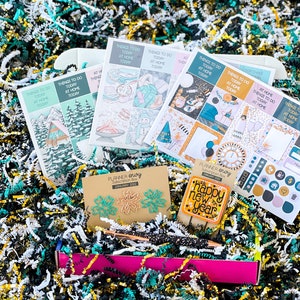 May include: A pink box filled with colorful paper shreds and planner accessories. The box contains a set of planner stickers with a New Year's theme, a set of paper clips with a floral design, a pen with gold glitter, and a bookmark with the text "Happy New Year".