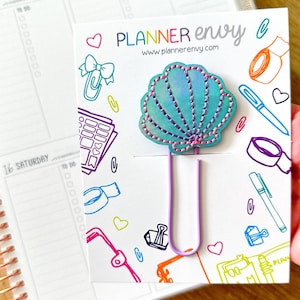 Puede incluir: Un clip en forma de concha con un diseño azul y rosa, sujeto a una tarjeta blanca con el texto "PLANNER envy" y garabatos coloridos. Un planificador está en el fondo.