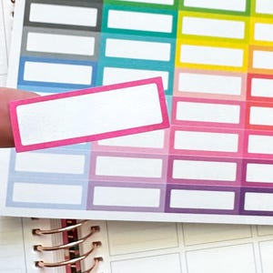 May include: A close-up of a sheet of planner stickers with a variety of colors and the text "PLANNER envy". A pink-bordered sticker is held up, showing a white rectangle. The stickers are on a spiral-bound planner.