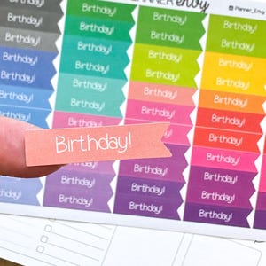 May include: A sheet of colorful planner stickers with the word "Birthday!" in white. The stickers are in various colors, including pink, purple, green, and yellow. A single sticker is held up by a finger.