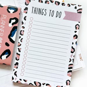 May include: A white Things To Do notepad with a pink and black leopard print border. The notepad has a pink banner and a list of circles for check-off. A matching planner is visible in the background.