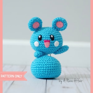 May include: A crocheted blue and pink mouse amigurumi toy with a smiling face. The toy is standing on a white wooden surface. The text "PATTERN ONLY" is visible in the lower left corner of the image. The text "by A Hare Affair" is visible in the lower right corner of the image.