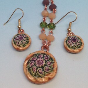 May include: A copper chain necklace with a large, round pendant featuring a floral design in shades of green, pink, and brown. The pendant is attached to the chain with a copper wire. The necklace comes with matching earrings with smaller versions of the pendant.