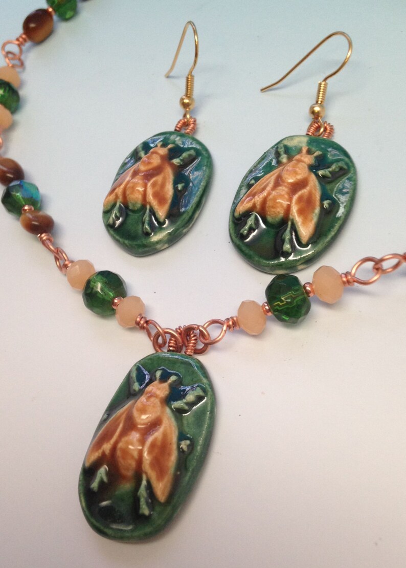 May include: A green and brown ceramic bee pendant necklace with matching earrings. The necklace features copper wire and green and beige beads.