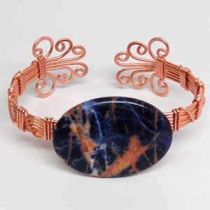 May include: A copper wire wrapped bracelet featuring a blue and orange oval gemstone. The bracelet has decorative swirls at each end.