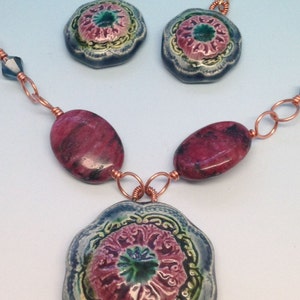 May include: A necklace with a large, blue and green floral pendant, two matching earrings, and pink and black oval beads. The necklace is made with copper wire.