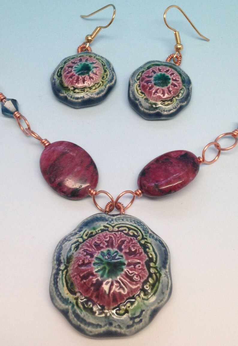 May include: A necklace with a large, blue and green floral pendant, two matching earrings, and pink and black oval beads. The necklace is made with copper wire.