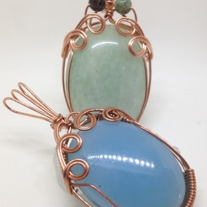 May include: Two wire-wrapped pendants with oval-shaped stones. The top pendant has a light green stone and copper wire with a dark green and black bead at the top. The bottom pendant has a light blue stone and copper wire.