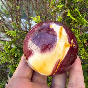 May include: A polished spherical stone featuring a smooth surface with swirling patterns of deep maroon, burgundy, and creamy yellow. The orb displays natural mineral inclusions and a glossy finish that reflects light across its rounded form.