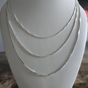 May include: Three delicate silver box chain necklaces displayed on a white bust. The necklaces are of varying lengths, creating a layered effect. The chains have a subtle shine, reflecting light.