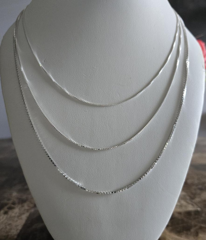 May include: Three delicate silver box chain necklaces displayed on a white bust. The necklaces are of varying lengths, creating a layered effect. The chains have a subtle shine, reflecting light.