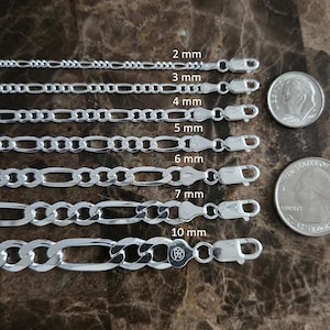 May include: Silver Figaro chain necklaces in various widths, from 2 mm to 10 mm, displayed on a brown surface. A dime and a quarter are included for size comparison. The chains have lobster claw clasps.