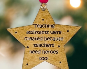 Może przedstawiać: Drewniana ozdoba w kształcie gwiazdy z wygrawerowanym tekstem: Teaching assistants were created because teachers need heroes too! Ozdoba zawieszona jest na sznurku zwieńczonym pojedynczym, okrągłym, jaskraworóżowym koralikiem.