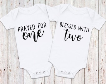 Może przedstawiać: Dwa białe body dla niemowląt z krótkimi rękawami. Body po lewej stronie ma napis "Prayed for one", a body po prawej stronie ma napis "Blessed with two".