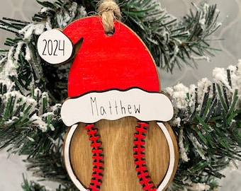 Może przedstawiać: Drewniana ozdoba w kształcie piłki do baseballa z czerwoną czapką Mikołaja, z rokiem 2024 na pomponie i imieniem Matthew na białym otoku. Piłka ma wykończenie z naturalnego drewna z czerwonymi detalami szwów i białą, okrągłą obwódką.